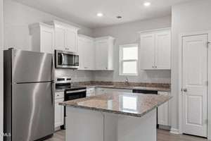 A modern kitchen with white cabinets and stainless steel appliances, including a refrigerator, oven, and microwave. A granite-topped island is centered, and there are recessed ceiling lights. A window provides natural light next to the sink.