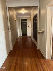 A narrow hallway with light-colored walls and white wainscoting features a wooden floor. The ceiling has a single light fixture. A built-in cabinet with glass doors is on one side. An open doorway at the end leads to another room.