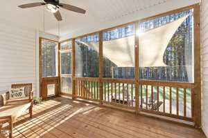 Enclosed porch with wooden floor and white ceiling. A ceiling fan is on the left, and a seating area with a chair and "Be Kind" pillow is visible. Large windows show a sunny deck outside, with a forest in the background. Sunlight streams in.