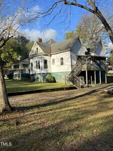 A large, two-story house with a white exterior and green base is surrounded by trees. It has a porch with stairs leading down to a grassy yard. The sky is clear with a few clouds.