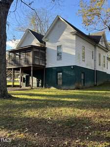 A two-story house with white siding and a green lower section is visible. It features a screened-in porch on the upper level. The yard is grassy with a few fallen leaves, and there is a tree in the foreground. The sky is clear and blue.
