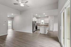 A modern, unfurnished kitchen and living area with light wood flooring. The kitchen has white cabinets, a stainless steel refrigerator, and a small island. The space features recessed lighting and sliding glass doors leading outside.