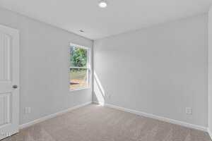 A small, empty room with light gray walls and beige carpet. A single window on the left wall lets in natural light. There is a white door on the left, and a ceiling light is on. The window shows a view of greenery outside.