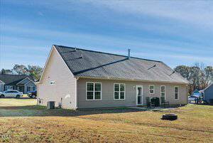 Single-story beige house with a sloped roof and multiple windows on a clear day. The back yard features a few patio chairs and a small fire pit on the grass. Neighboring houses and trees are visible in the background.