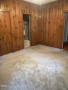 Room with wood-paneled walls and light-colored carpet. There is an open door leading to a bathroom with a visible shower. Two doorways are present, one to the left and one to the right. The room is empty and well-lit.