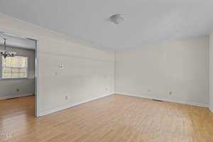 An empty room with light wood flooring and beige walls. A ceiling light fixture is centered in the room. An open doorway leads to another room with a window and a chandelier. Natural light enters through the windows.