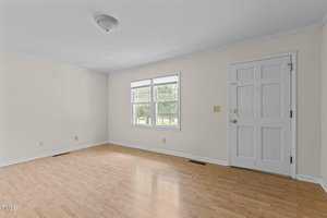 An empty room with light wood flooring and cream-colored walls. It has a window with blinds and a white door. A ceiling light fixture is centered in the room, and there are electrical outlets on the walls.