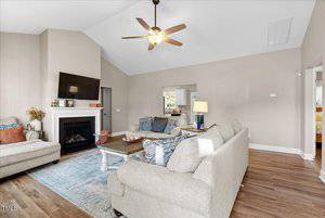 A living room with beige walls and wooden flooring features two beige sofas surrounding a wooden coffee table. A fireplace with a TV mounted above it is on one wall. A ceiling fan hangs from the vaulted ceiling, and a glimpse of a bedroom is visible.