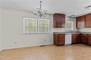 A kitchen with wooden cabinets, a marble countertop, and a stainless steel dishwasher. There's a two-window setup with blinds, a double sink, and a ceiling-mounted light fixture. The floor has a wooden finish, and the walls are light-colored.