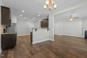 Spacious open-plan kitchen and living area with wooden floors. The kitchen features dark cabinetry, stainless steel appliances, and a central island with a granite countertop. A ceiling fan and a fireplace are visible in the adjoining living space.