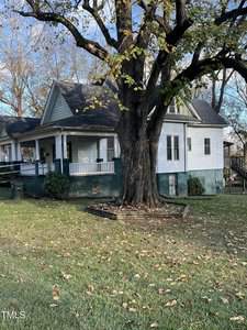 A white house with a porch and a gabled roof stands behind a large tree with thick branches. The yard is covered in grass and scattered leaves. A trash bin is visible near the porch, and a wooden retaining structure surrounds the tree.