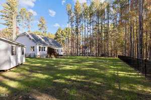 A single-story white house with a porch is surrounded by a grassy yard. A black fence borders one side. Trees with autumn leaves line the backyard, and a small shed is visible on the left. The sky is clear with a few clouds.