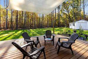 A wooden deck with four black Adirondack chairs arranged around a small round table. A sail shade is stretched above, and a grassy yard with a shed is in the background, surrounded by trees.