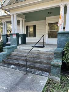 The image shows a front porch of a house with green and white exterior. There are concrete steps with a black handrail leading up to the porch. A "For Sale" sign is posted. The front door has a screen door and a flag is visible to the right.