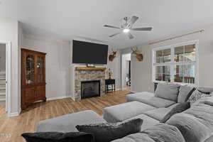 A living room with a large gray sectional sofa, a stone fireplace, and a flat-screen TV mounted above. Two deer head mounts are on the wall. There's a wooden cabinet on the left and a view of a dining area through a doorway. Ceiling fan overhead.