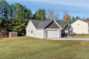 A suburban single-story house with a gray roof and beige siding is shown. It has a double garage, a small front porch, and a well-maintained lawn. A small shed is visible in the background, surrounded by trees under a clear blue sky.