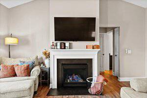 A living room with a beige couch and patterned cushions, a tall lamp, and a fireplace beneath a wall-mounted TV. A basket with blankets sits near the fireplace. Wooden flooring and light-colored walls complete the cozy setting.