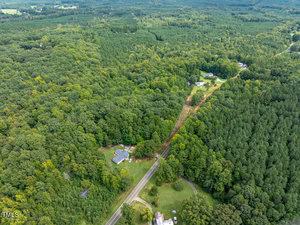 Aerial view of a dense, green forest with a rural road cutting through it. Several houses are scattered near the road, surrounded by trees. The landscape is lush and expansive, with no other structures visible in the distance.