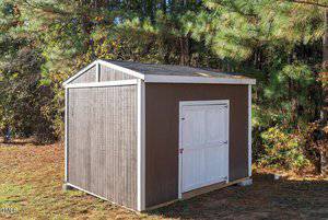 A brown shed with a white door and a gabled roof is situated on a grassy area. It is surrounded by trees, with some sunlight filtering through the branches. The ground is partially covered with fallen leaves.