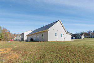 A single-story, light-colored house with a sloped roof is situated on a grassy lawn. The sky is clear and blue. There are trees in the background, and neighboring houses are visible on either side.