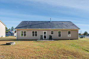 A single-story beige house with a dark roof is shown, featuring multiple windows and a small patio with two chairs in the back. The house sits on a grass-covered yard under a clear blue sky.