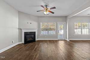 A spacious living room with light gray walls, a wooden floor, and a ceiling fan. A fireplace is on the left. Two large windows and a glass door on the right side allow natural light to enter the room.