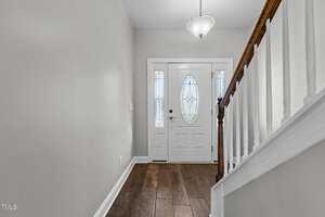 A hallway featuring light green walls, a wooden stair rail, and a dark wood floor. The white front door has an oval glass panel with decorative patterns, and a hanging pendant light illuminates the area.