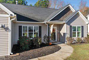 A single-story house at 315 Keeneland Drive features gray siding, a black shingled roof, and white-trimmed windows. The front yard in Oxford showcases simple landscaping with small shrubs and a pumpkin by the door. A flag and an "AUTUMN" sign add charm near the entrance.