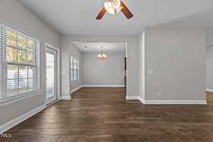 An empty room with light gray walls and wooden flooring. A ceiling fan with wooden blades hangs above. Sunlight comes in through large windows and a glass door. A chandelier is visible in an adjoining room.