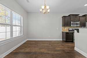 A kitchen and dining area with wooden flooring and light gray walls. The kitchen features dark brown cabinets, a stove, microwave, and a tile backsplash. A window with blinds is on the left, and a ceiling light fixture hangs above.