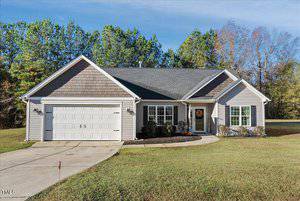 A single-story house with gray siding and a two-car garage. It has a gabled roof and a front porch with a decorative wreath. There are trees in the background and the lawn is neatly maintained.