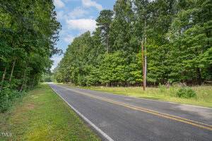 A paved road stretches through a lush forest with tall trees on both sides. The sky is partly cloudy, and sunlight filters through the leaves. A grassy shoulder runs alongside the road.