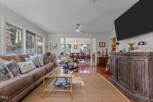 A cozy living room with a large beige sofa, decorative pillows, and a glass coffee table on a woven rug. A flat-screen TV is mounted above a wooden cabinet. The room has large windows and a view into a dining area with a wooden floor.
