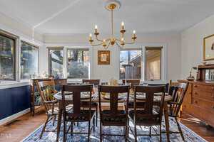 A dining room with a wooden table set for eight, featuring matching chairs. A brass chandelier hangs above. The room has large windows, a sideboard, and framed art on beige walls. A patterned rug covers the wooden floor.