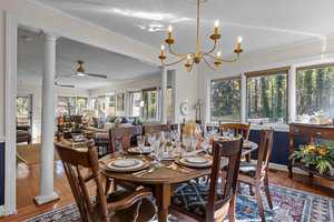 Dining room with a set table featuring elegant dinnerware and glassware. A chandelier hangs above. The room opens into a living area with sofas and a large window view of trees outside. A wooden sideboard is decorated with a floral arrangement.