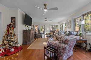 A cozy living room with wood flooring features a decorated Christmas tree, a large wall-mounted TV, and a collection of sofas and armchairs. The space is well-lit with large windows and two ceiling fans. A rug and various plants add warmth to the room.