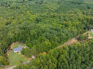 Aerial view of a dense forest with a small clearing containing a house and a yard. A road runs alongside the forest, and another structure is partially visible in the distance. The trees are lush and green.