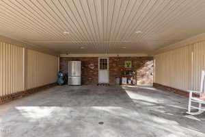 A carport with a concrete floor, beige ceiling, and walls. A brick wall at the back has a white door and a small window. A refrigerator, bags, and decorations are to the left. A rocking chair with a cushion is to the right. Shadows are cast on the floor.