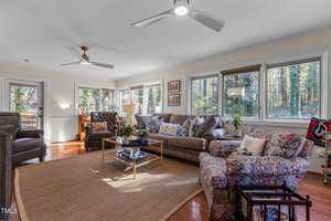 A cozy living room with large windows, beige walls, and wood flooring. It features a brown sectional sofa, patterned armchair, ceiling fans, and a woven rug. The room has decor items, plants, and a view of trees outside.