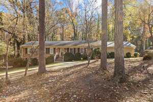 A single-story brick house with a shingled roof is surrounded by tall trees and shrubs. The driveway leads to the front, and autumn leaves cover the ground. The background features a wooded area under a clear blue sky.