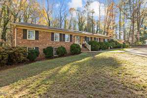 Nestled in a wooded area with autumn trees, the single-story brick house at 161 Pine Cone Drive features multiple windows and a welcoming porch. Its lawn, partially covered with grass, complements the curved driveway on this clear blue sky day in Oxford.