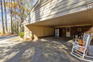 A covered carport attached to a house with a brick wall features a white rocking chair, pumpkins, and an outdoor fridge. Trees and a paved driveway are visible in the background under a clear sky.