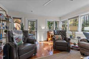 A sunlit living room features two brown leather armchairs and a bookshelf on the left. Large windows offer a view of trees outside. A small desk with a chair is placed near the window. A floor lamp and decor items add to the cozy atmosphere.