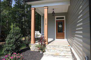 Front porch of a house with a brown door and a small window displaying a welcome sign. The porch has a white bench on the left and the number "3613" on a wooden column. There are plants and flowers in a landscaped area beside the steps.