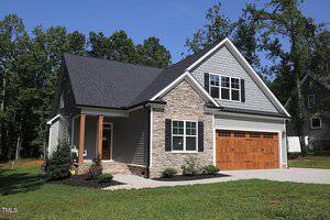 Located at 3613 River Watch Lane in Franklinton, this modern house features a gray and stone exterior with a steep gabled roof and multiple windows. The property boasts a well-maintained lawn with shrubs and a driveway leading to a wooden garage door, all framed by trees in the background.