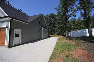 Side view of a modern house with gray siding, featuring a wooden garage door and a concrete driveway. There is a white fence on the right and a backdrop of green trees under a clear blue sky.