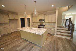 A modern kitchen features light-colored cabinets and a large island with a marble countertop. Stainless steel appliances, including an oven and microwave, are visible. Overhead pendant lights illuminate the space. Stairs are seen on the right.