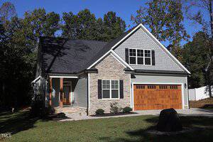 A modern house in a wooded area with a gray exterior and stone accents. It has a pitched roof and a two-car garage with wooden doors. A lawn surrounds the house, and a boulder is visible in the foreground.