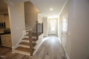 A modern hallway with light wood flooring leads to a staircase with wooden steps and dark railings. White walls and a white door at the end of the hallway create a bright and airy atmosphere. The kitchen is partially visible on the left side.