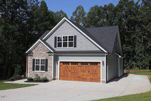 A two-story house with light gray siding and stone accents. It features a double garage with wooden doors and a dark gray roof. The house is surrounded by trees and has a concrete driveway leading up to it.